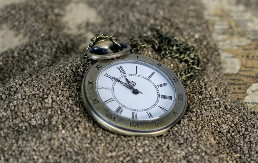 The picture shows a pocket watch that lies in sand. On the right hand side appears to be a part of a world map.