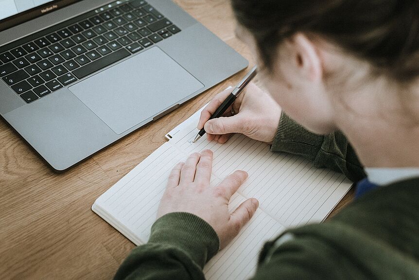 This picture shows a notebook that sits on a desk and a woman from the back who is writing into a book.
