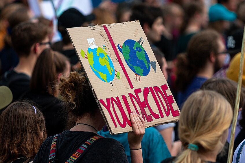 This picture shows a demonstration agains climate change. In the foreground there is a poster that shows a healthy earth and an ill one. In big letters one can read You Decide.