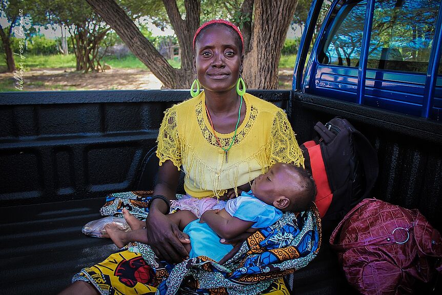 The picture shows a PoC mom who holds her sleeping son on her lap. Both sit on a lorry.